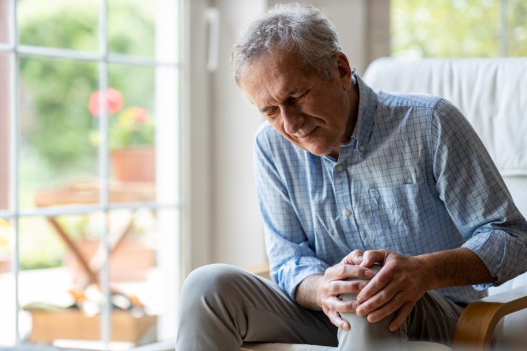 An older man sitting down on the couch holding his knee because he is in pain.